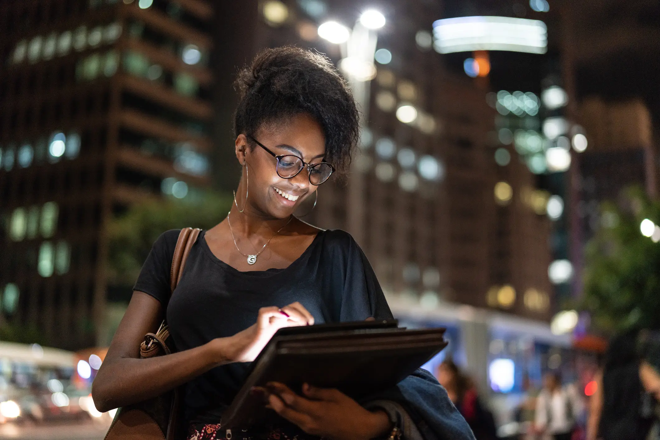 Mulher negra sorridente usando tablet à noite em ambiente urbano