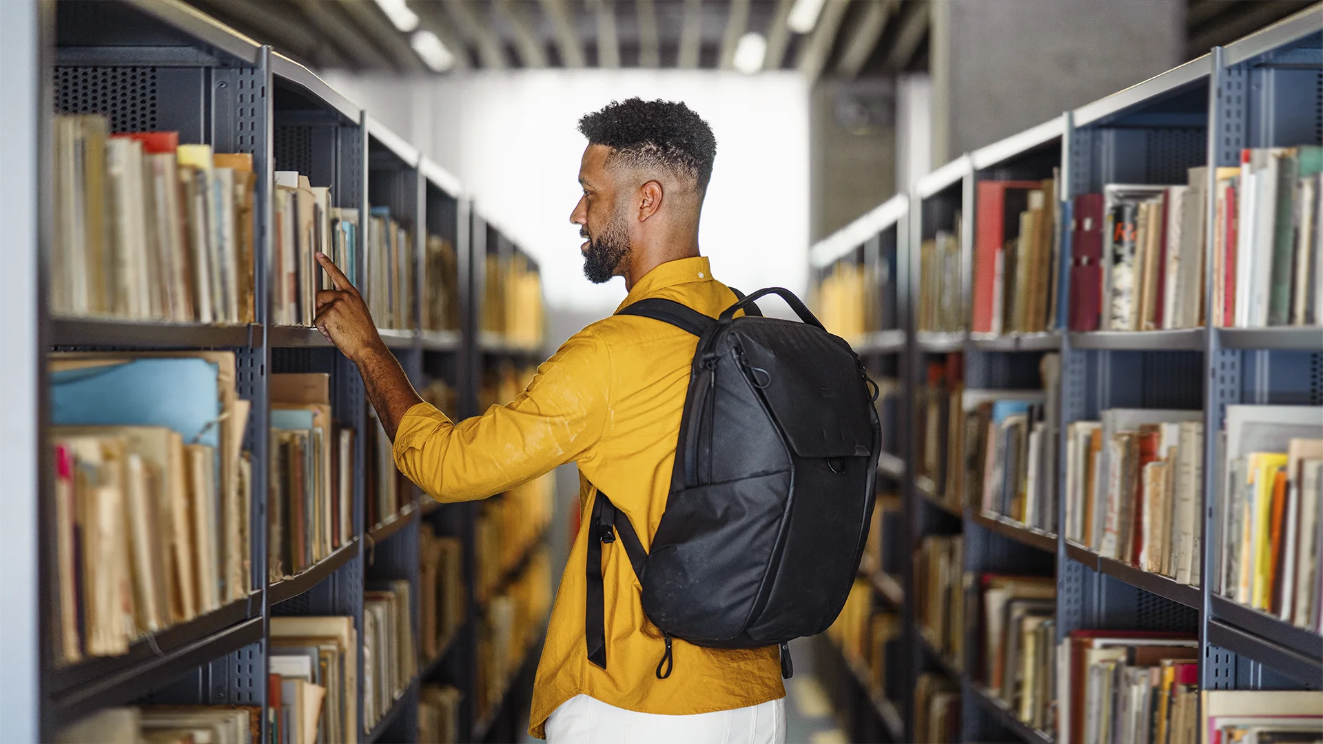Estudante universitário com mochila escolhendo um livro em uma estante de biblioteca