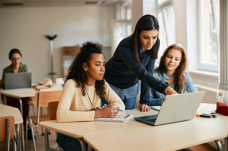 Professora auxiliando estudantes durante aula colaborativa com uso de tecnologia e computadores em ambiente acadêmico