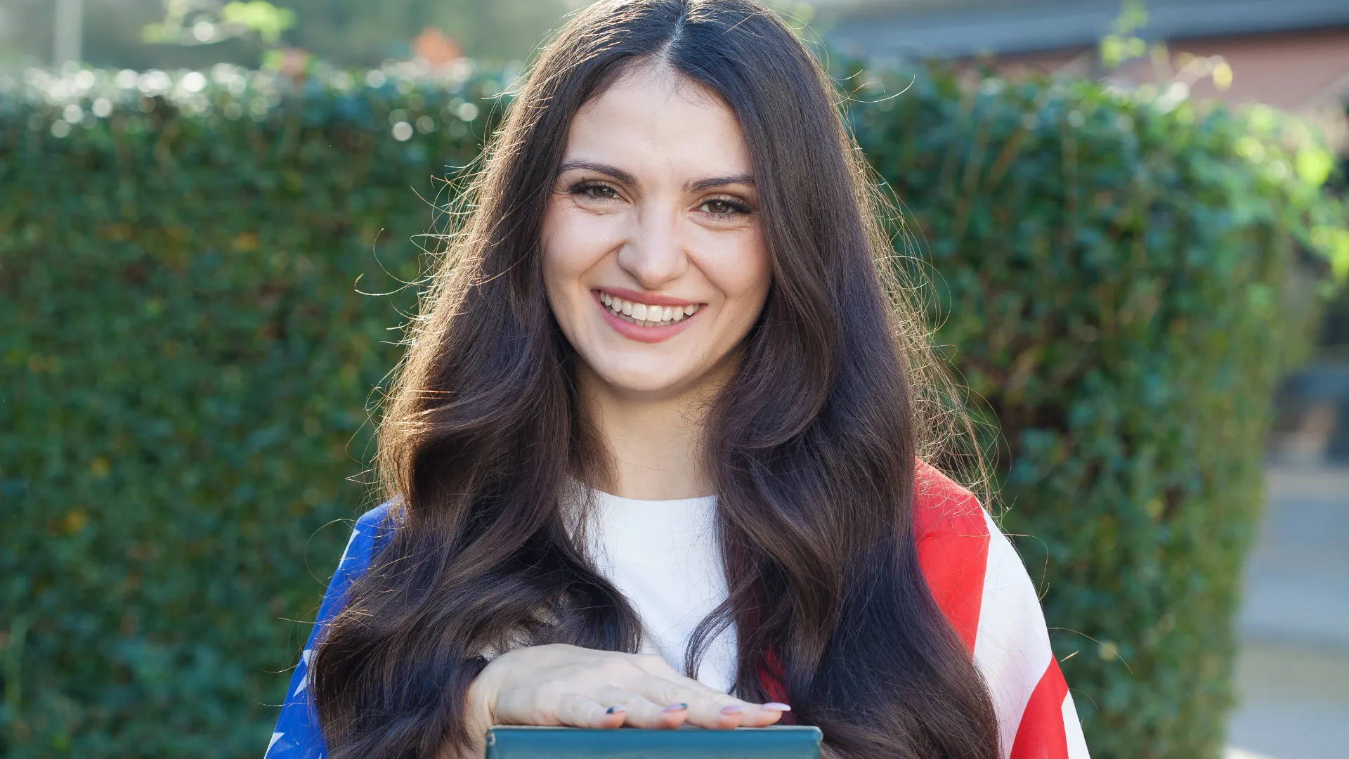 Jovem sorridente com cabelos longos e escuros usando uma bandeira dos Estados Unidos nos ombros