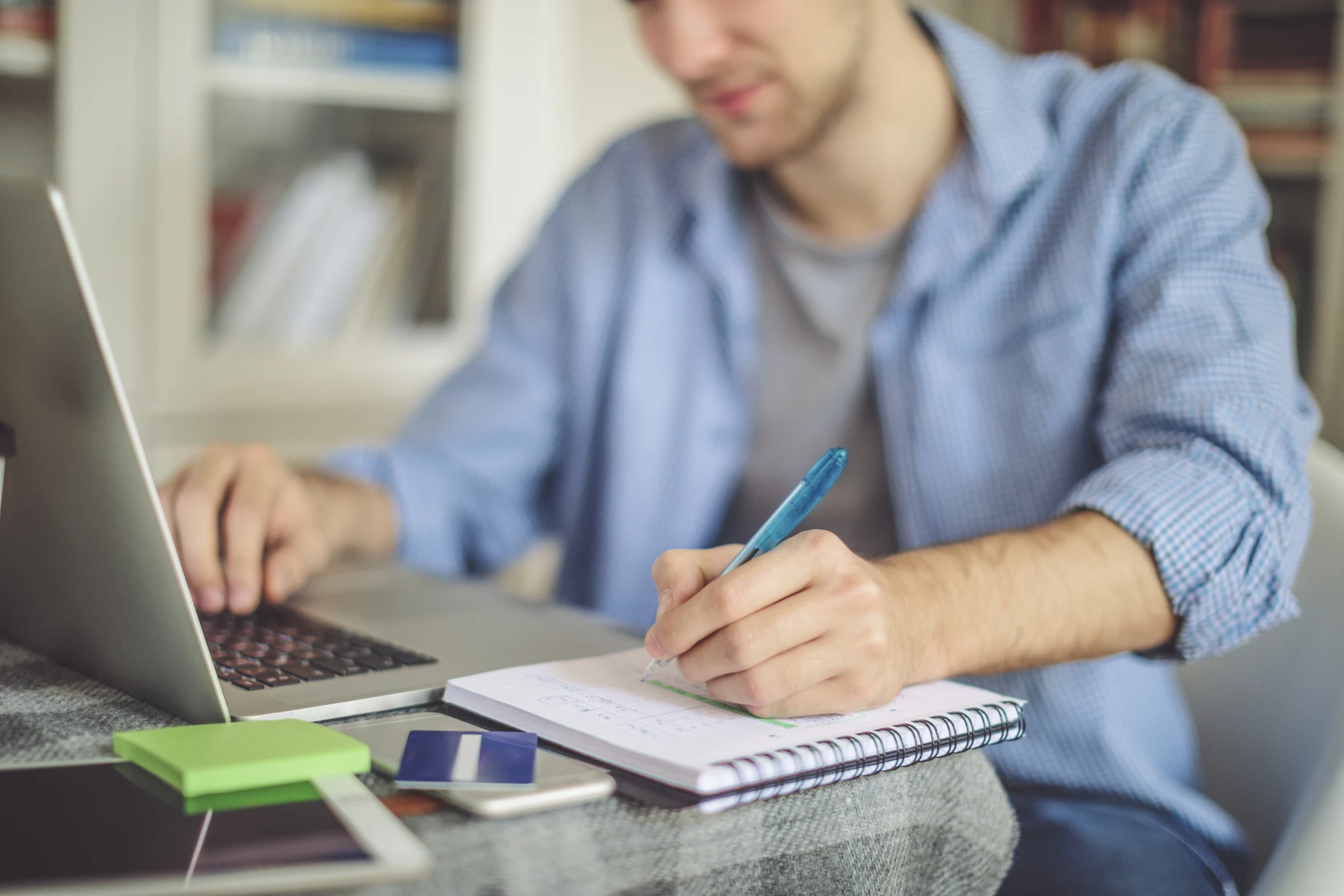 Estudante ou profissional escrevendo em um caderno enquanto usa um notebook em um ambiente de estudo ou home office.