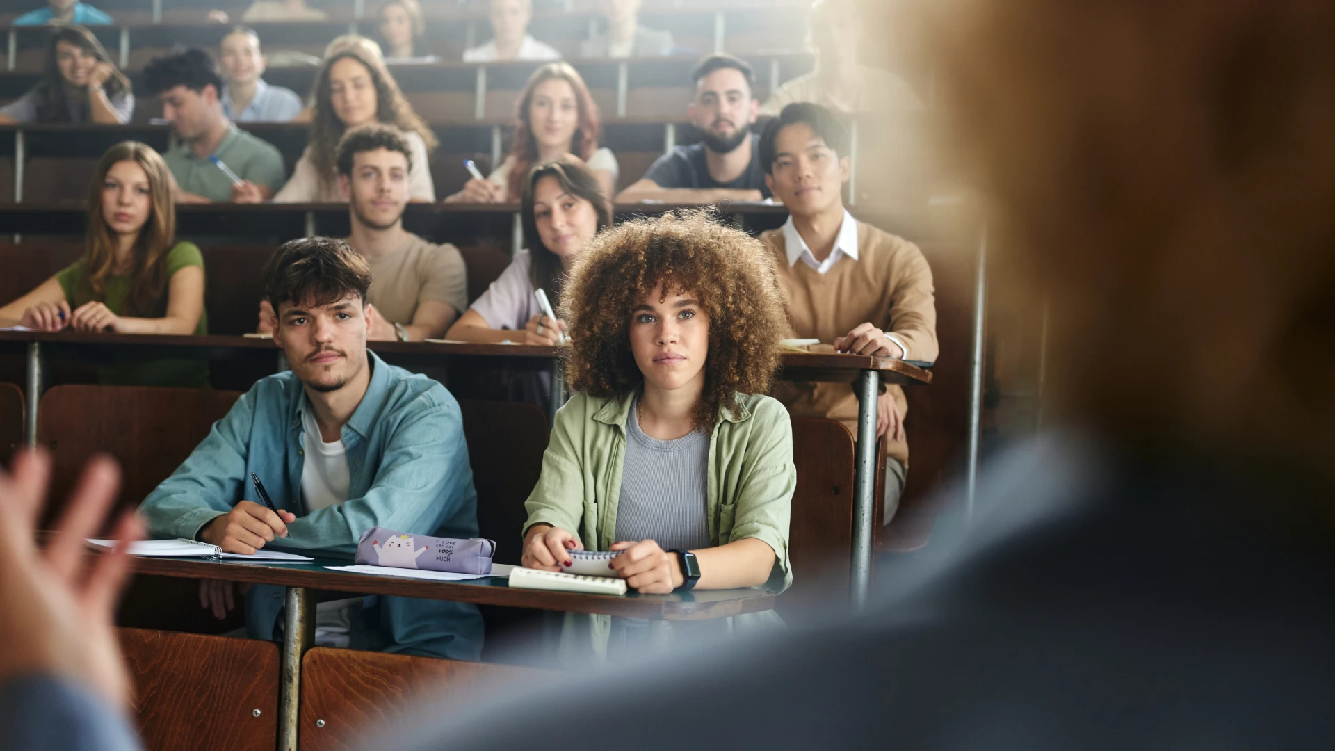 Grupo de estudantes diversos prestando atenção em uma aula presencial no ambiente universitário
