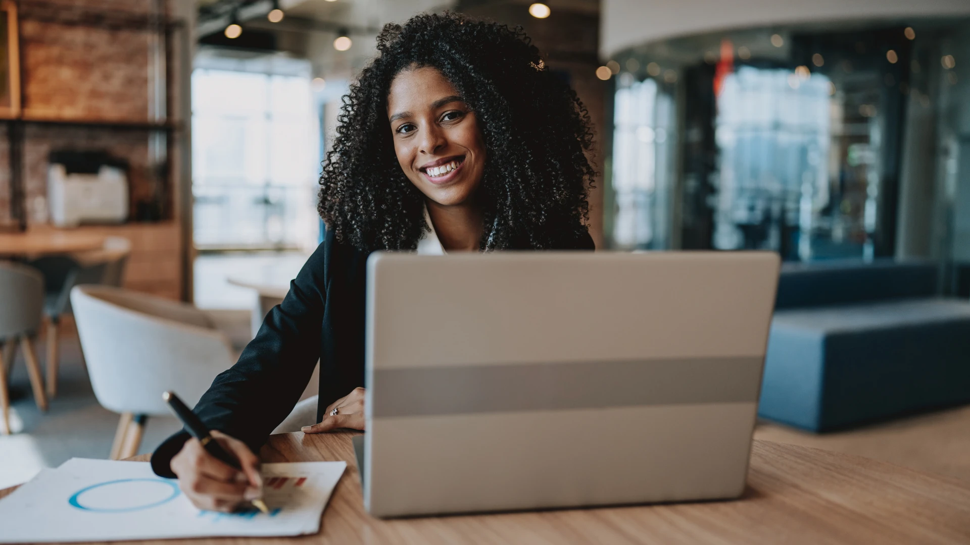 Mulher profissional negra sorridente trabalhando em um escritório moderno com notebook e documentos