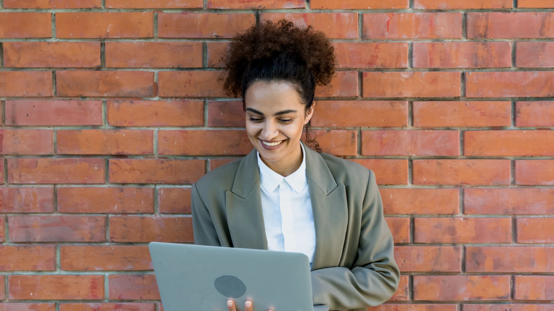 Mulher profissional sorrindo enquanto usa um laptop encostada em uma parede de tijolos