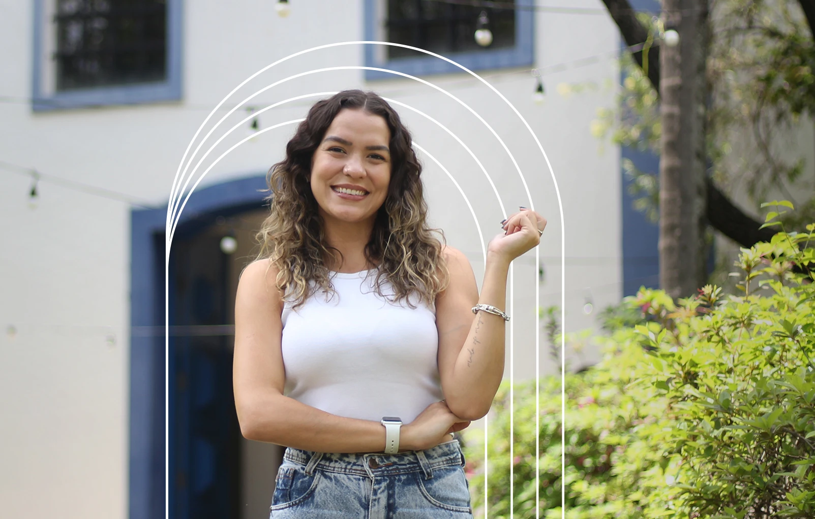 Jovem mulher sorridente com cabelos cacheados em um ambiente externo com fundo de arquitetura e vegetação