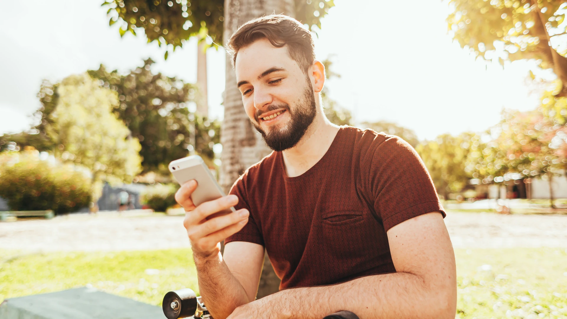 Jovem barbudo sorrindo enquanto usa um smartphone em um parque ensolarado