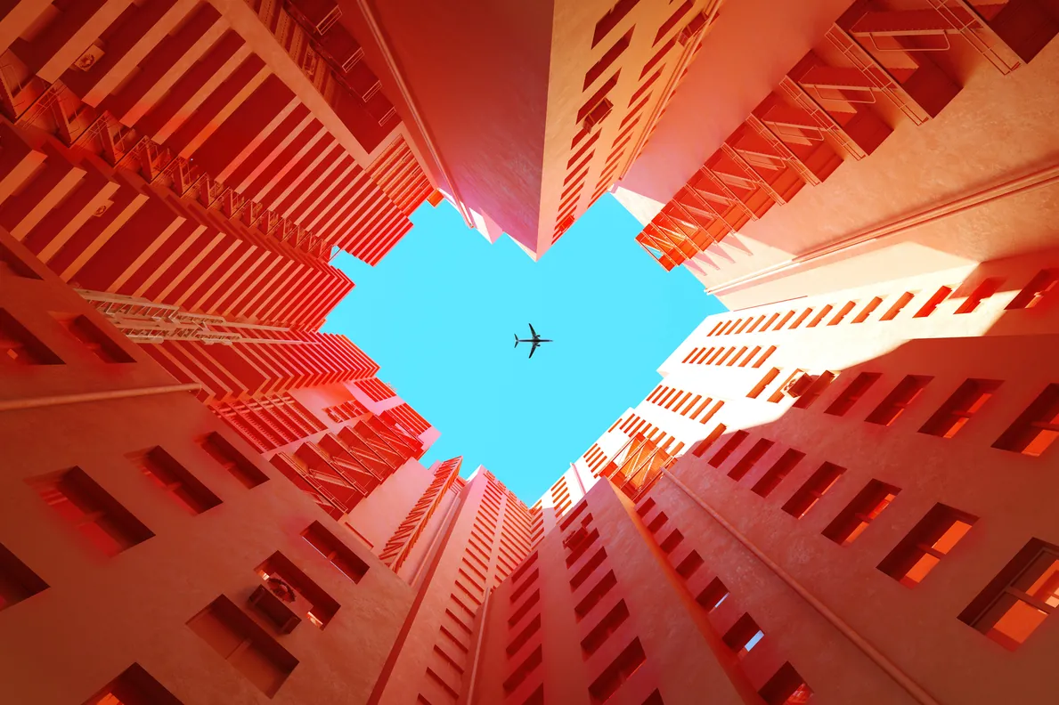Fotografia artística de um avião sobrevoando edifícios simétricos com tons alaranjados criando um efeito visual de coração no céu