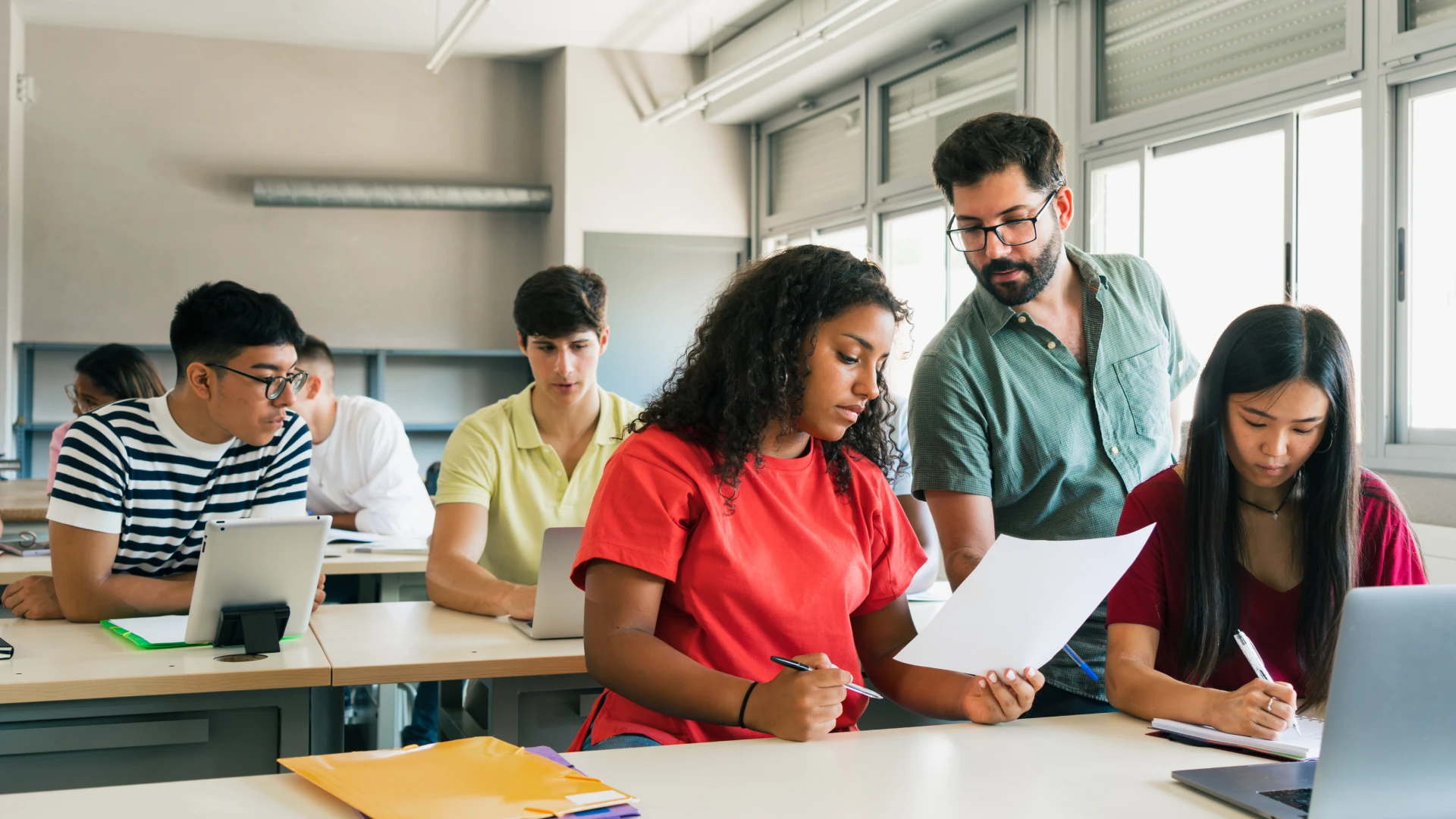 Professor universitário auxiliando alunos durante atividades práticas em sala de aula moderna com uso de tecnologia