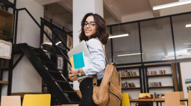 Estudante universitária sorridente segurando notebook e livros em uma biblioteca moderna
