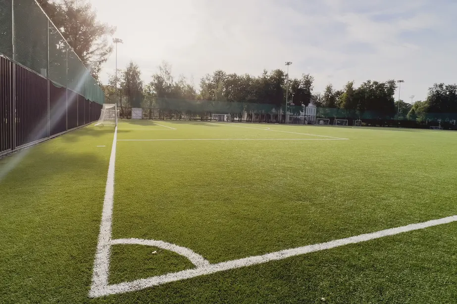 Campo de futebol sintético vazio em dia ensolarado. Vista ampla do gramado verde com marcações brancas, gol ao fundo, cercado com tela e árvores, iluminado por forte luz solar com reflexo