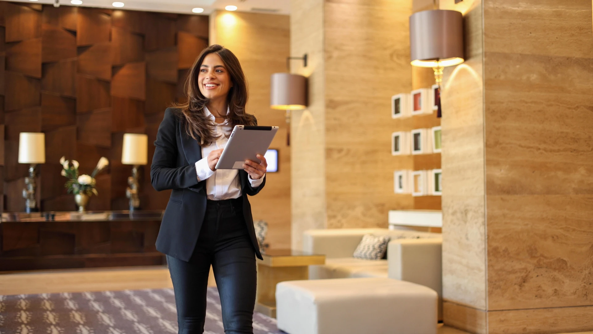 Mulher de negócios sorridente caminhando em um saguão elegante de hotel enquanto usa um tablet.