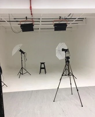 Estúdio fotográfico do Unifacs Santa Mônica com fundo branco infinito, dois guarda-chuvas de iluminação, tripés profissionais e um banco preto central. Espaço equipado para práticas de fotografia e produção audiovisual, tour virtual Professor Barros
