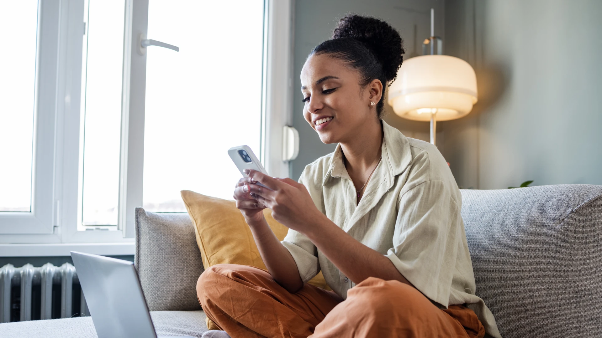 Jovem mulher negra sorridente usando smartphone sentada no sofá com um notebook