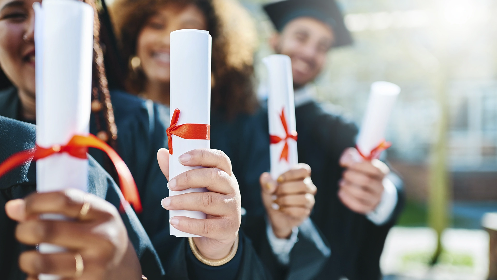 Grupo de formandos segurando diplomas enrolados com fitas vermelhas durante a cerimônia de graduação