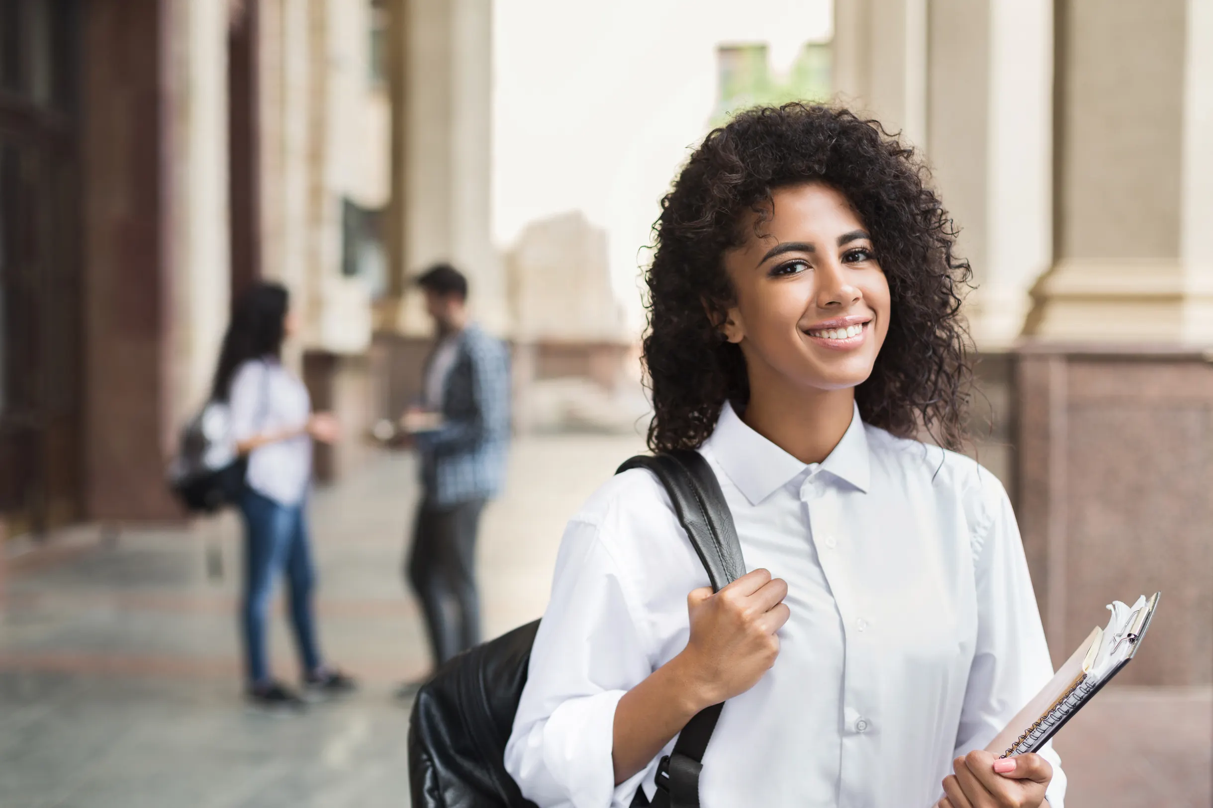 Estudante universitária sorrindo com mochila e caderno no campus da faculdade