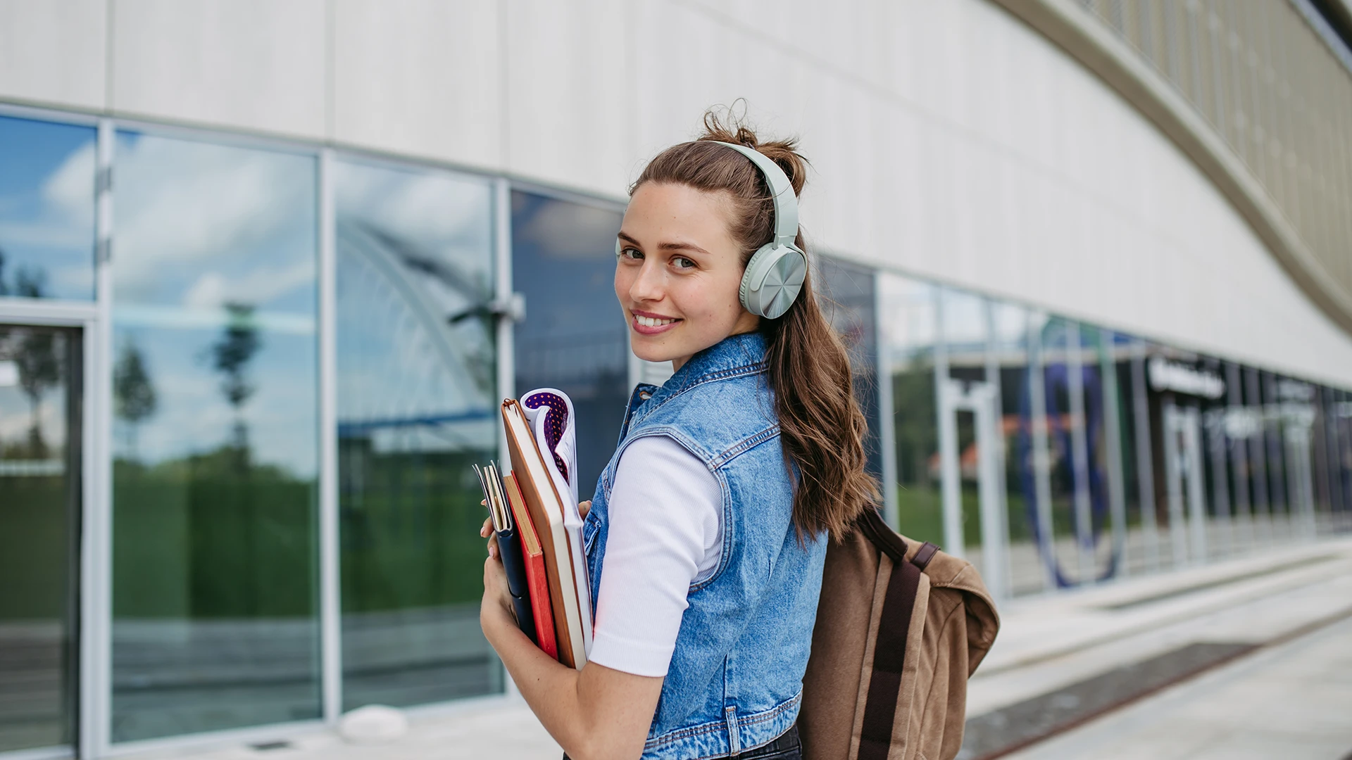 Estudante universitária sorridente segurando livros e usando fones de ouvido no campus