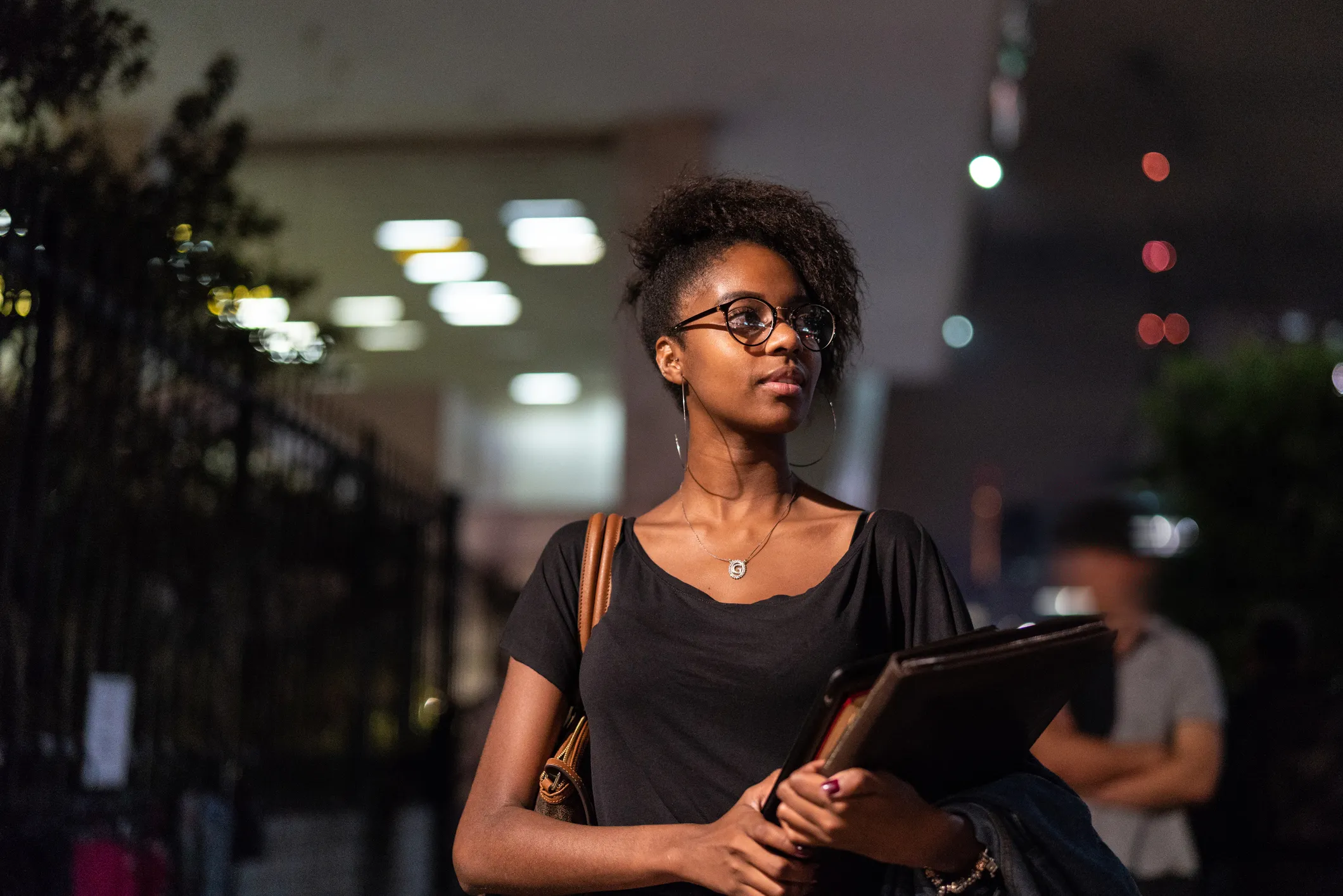 Estudante universitária negra de óculos segurando pastas à noite em campus