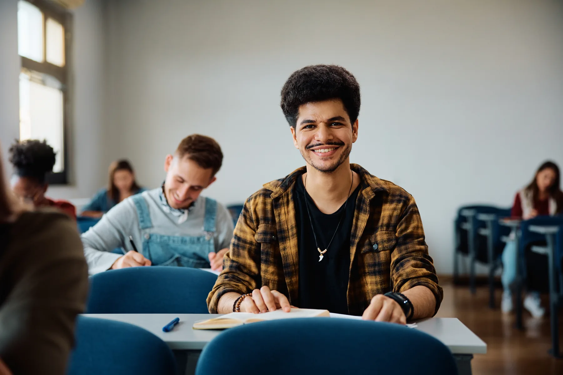 Estudante universitário sorridente sentado em uma sala de aula com outros alunos ao fundo