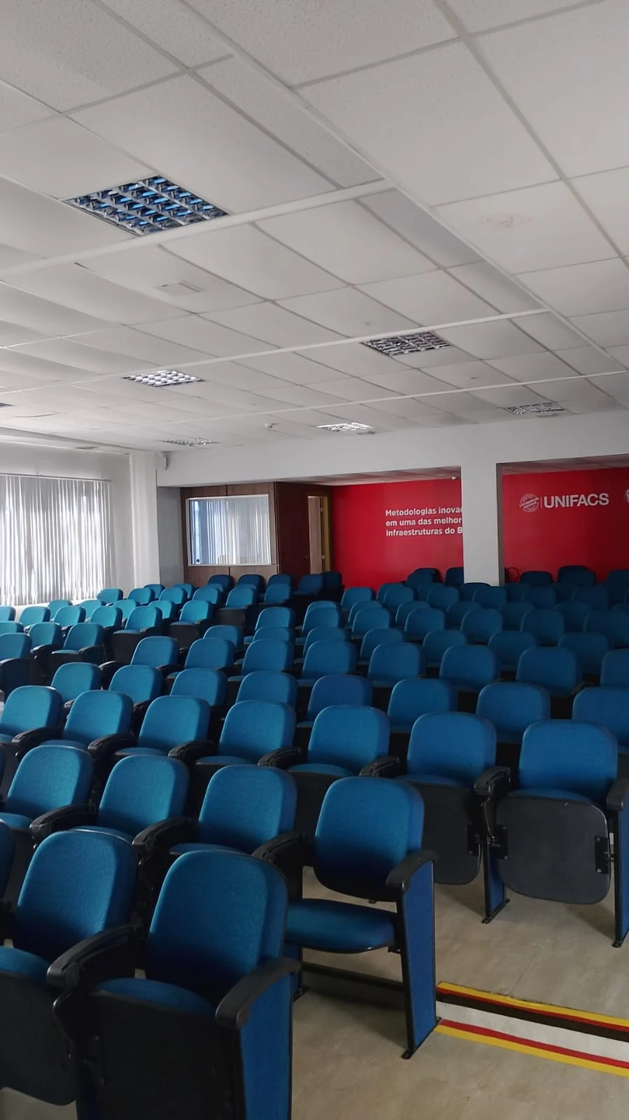 "Auditório do Unifacs com fileiras de cadeiras azuis e parede vermelha ao fundo com a logo Unifacs. Espaço amplo e moderno para palestras e aulas magna, parte do tour virtual conduzido pelo Professor Barros."