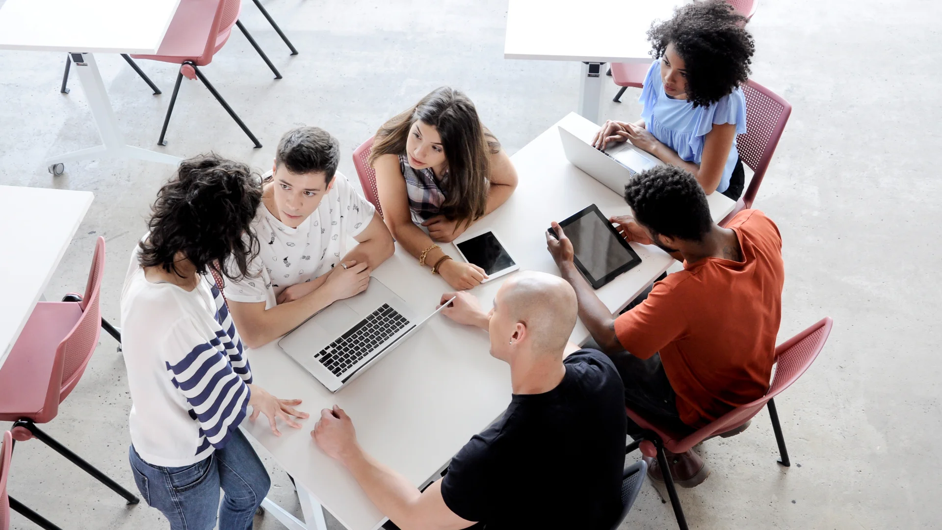 Grupo de estudantes universitários diversos colaborando em projeto com laptops e tablets em uma sala de aula moderna