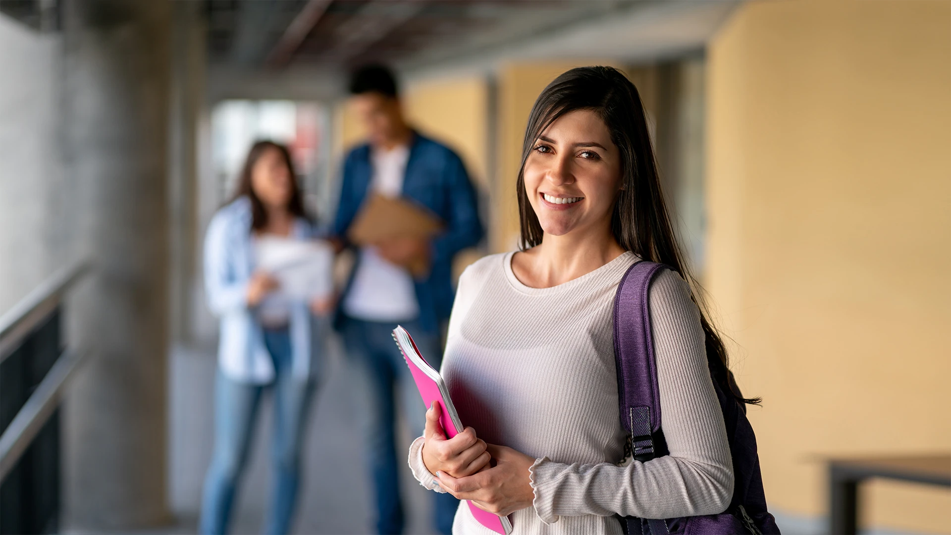 Estudante universitária sorridente segurando cadernos em um campus com outros alunos ao fundo
