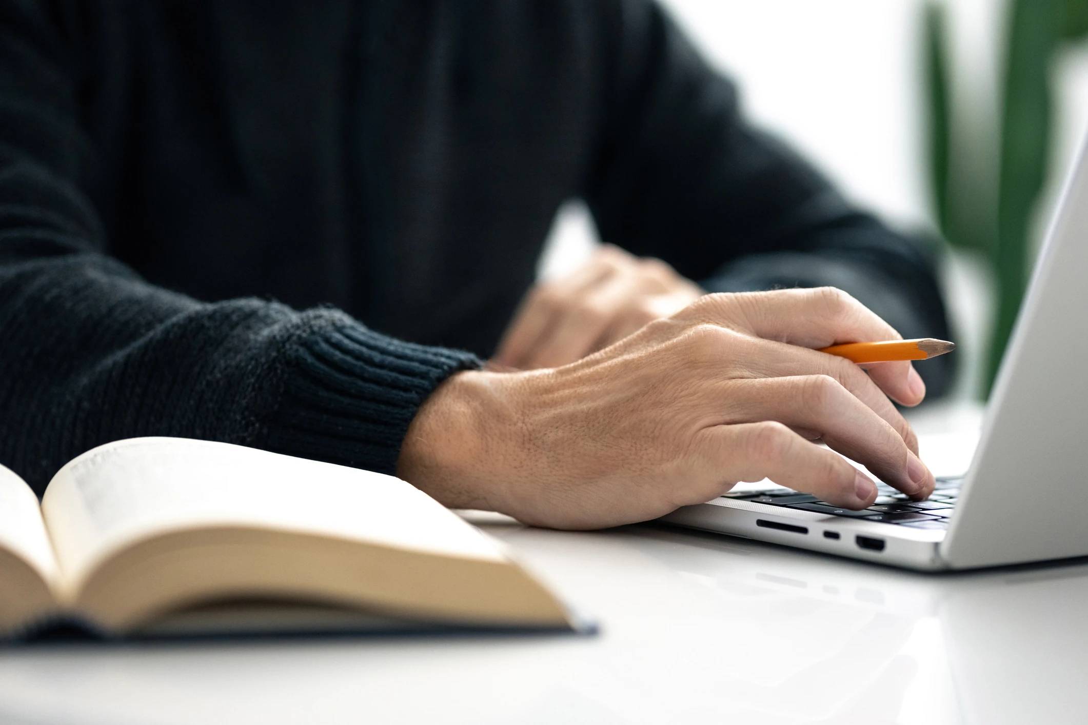 Estudante usando laptop com um livro aberto ao lado para pesquisa acadêmica