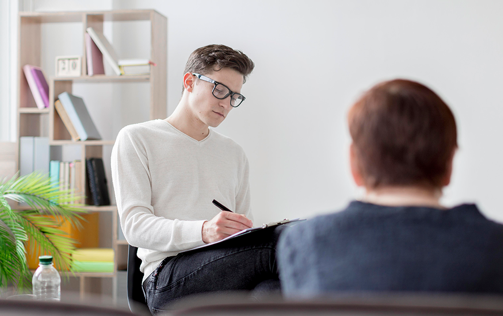 Psicólogo profissional realizando atendimento terapêutico e fazendo anotações em consultório acolhedor Psicólogo jovem de óculos fazendo anotações durante uma sessão de terapia com paciente