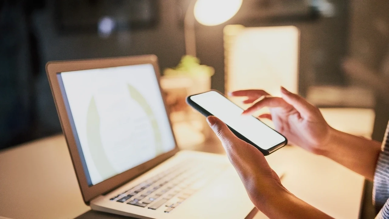Pessoa utilizando smartphone com tela em branco ao lado de um notebook aberto em ambiente de trabalho.