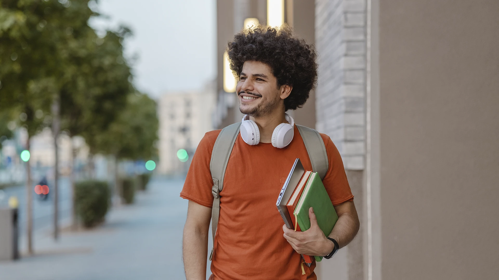 Estudante universitário sorridente com mochila, fones de ouvido e livros caminhando pelo campus