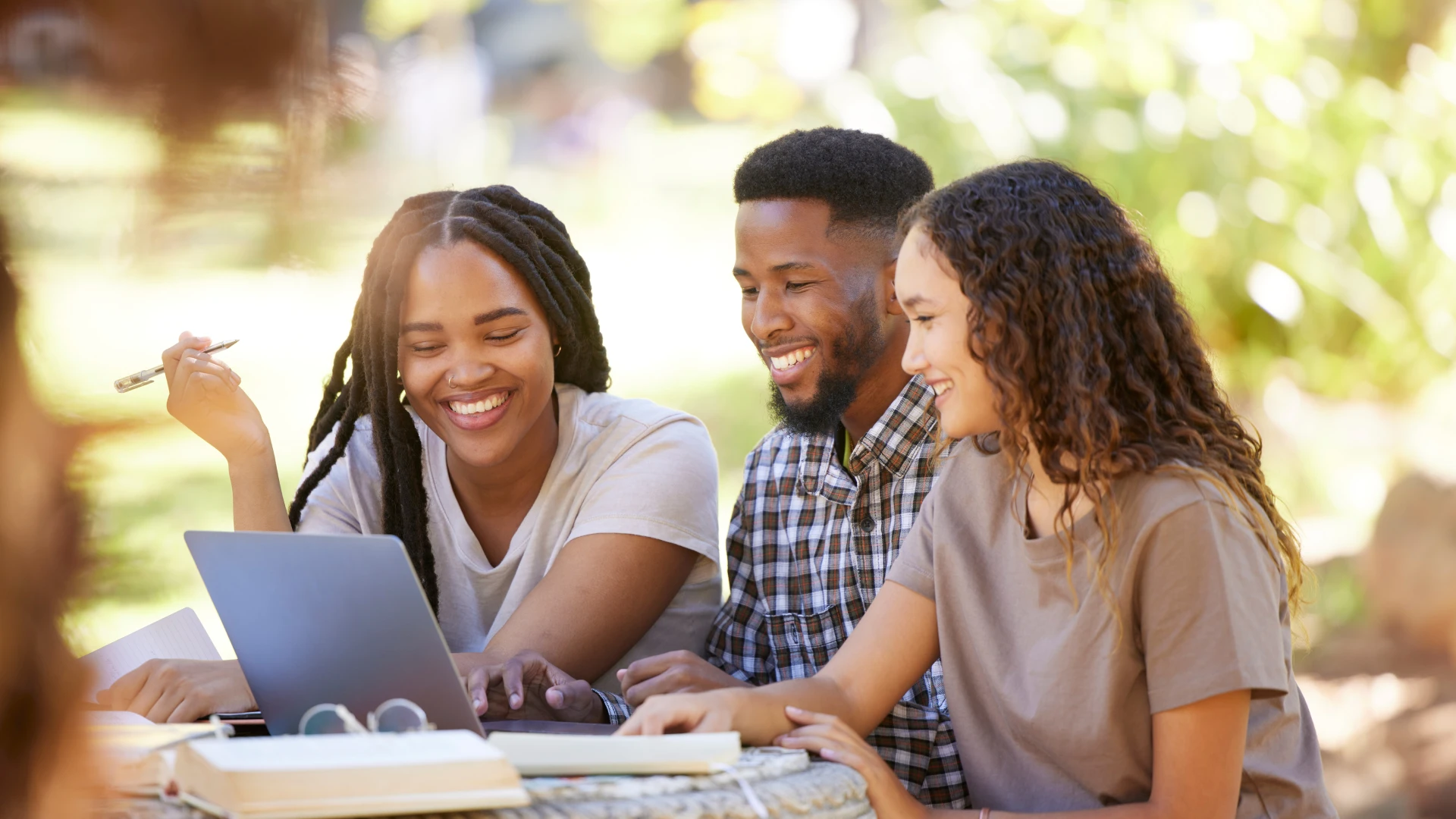 Estudantes universitários felizes colaborando em um projeto acadêmico usando um laptop em um ambiente externo ensolarado