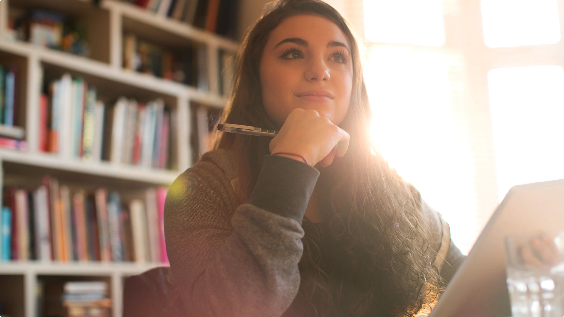 Estudante pensativa segurando uma caneta em uma biblioteca com luz solar