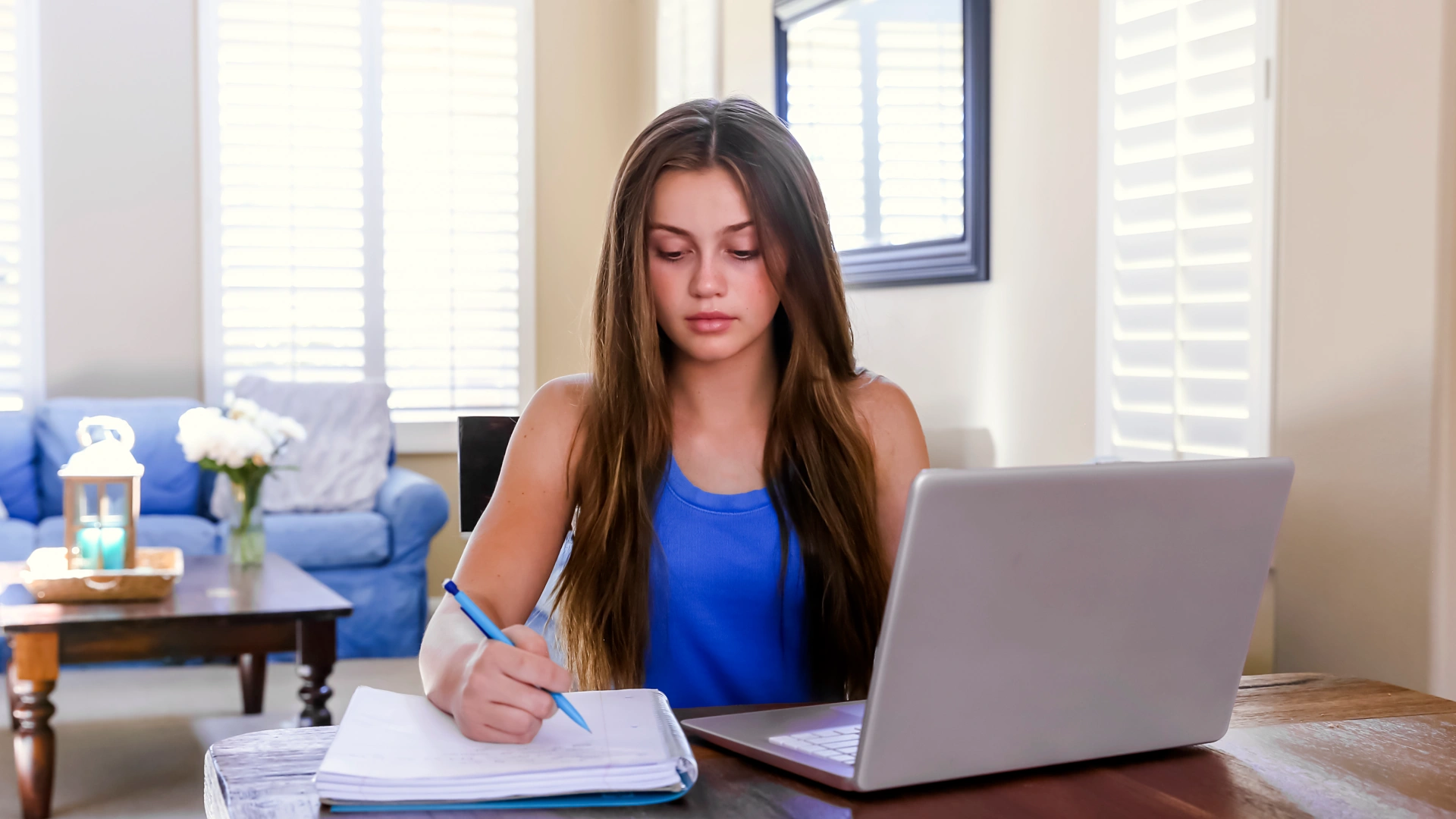 Estudante jovem concentrada escrevendo em um caderno enquanto utiliza um notebook em casa