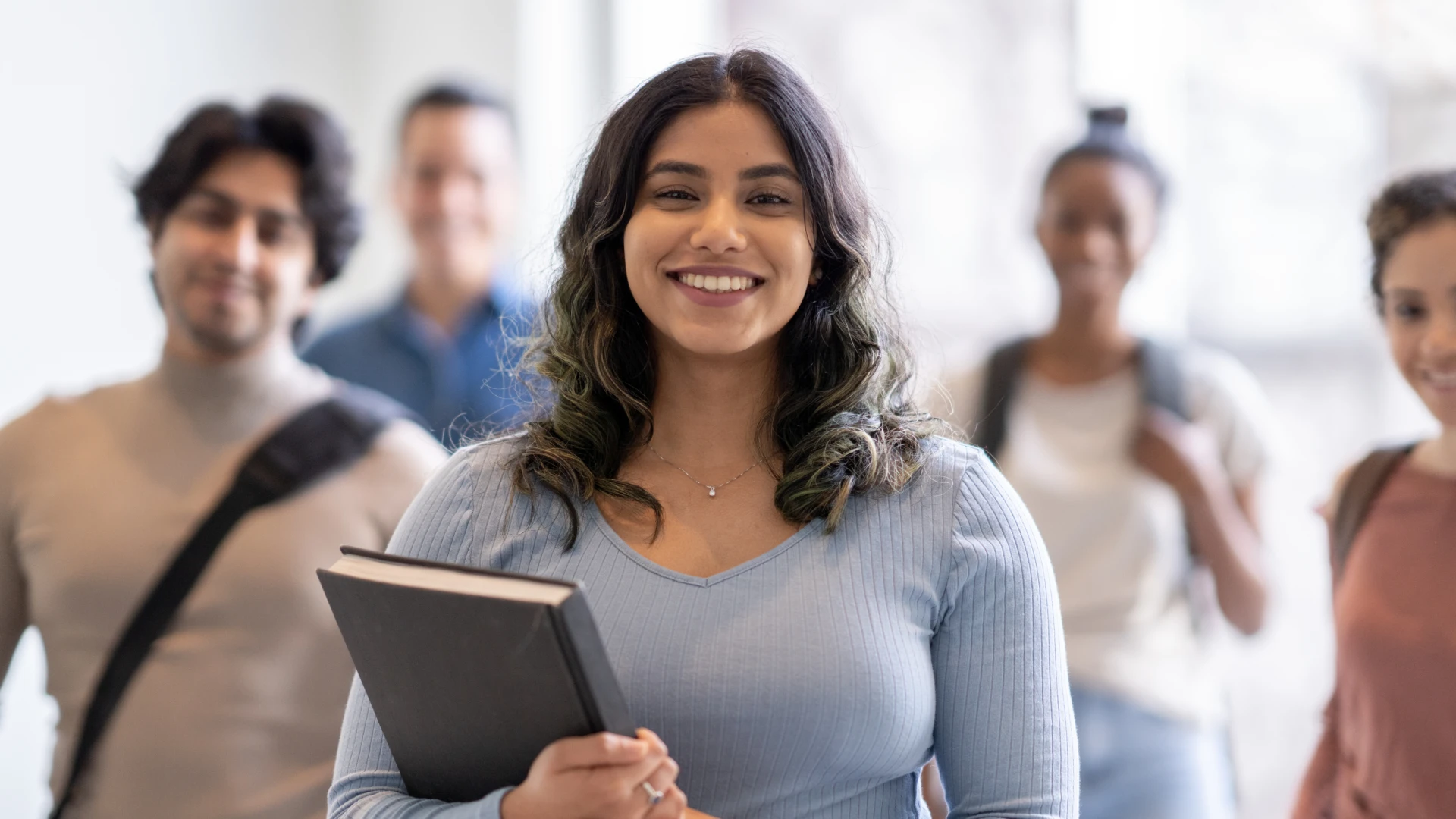 Estudante universitária sorridente segurando um livro com colegas ao fundo no campus