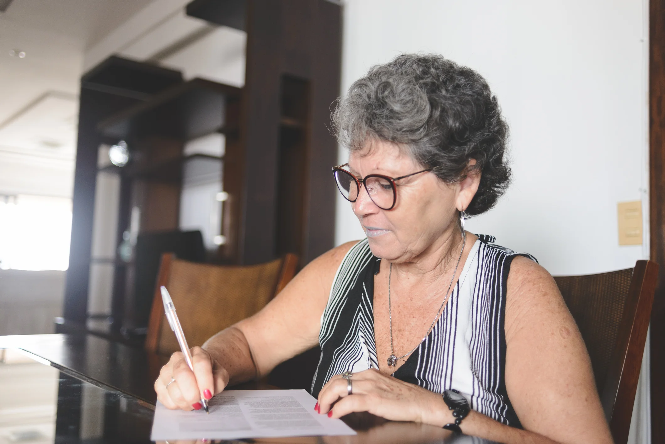 Mulher idosa de óculos escrevendo em uma folha de papel sentada à mesa