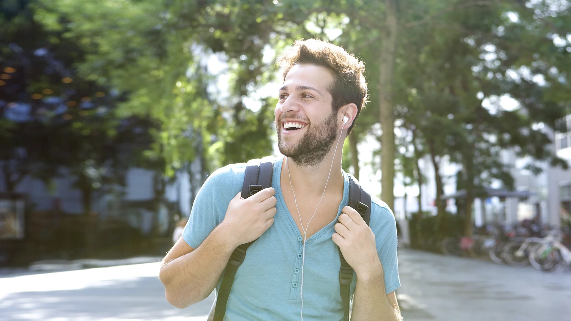 Estudante universitário jovem e sorridente caminhando ao ar livre com uma mochila e fones de ouvido.