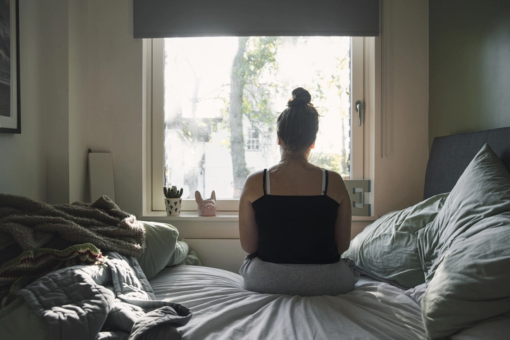 Mulher sentada na cama olhando pela janela do quarto durante o dia