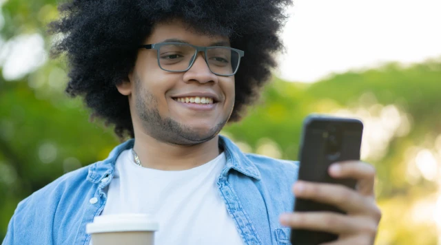 Homem jovem e sorridente com cabelo black power usando óculos e segurando um smartphone ao ar livre