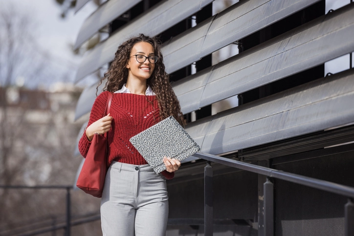 Estudante universitária sorridente caminhando no campus com cadernos e bolsa