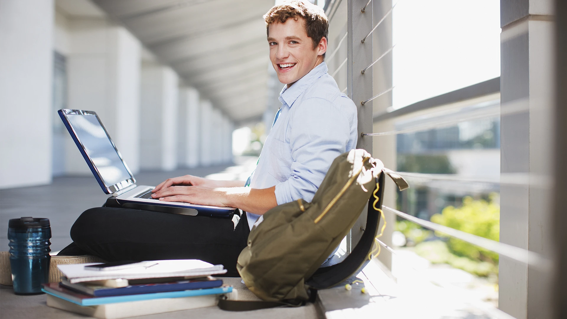 Estudante universitário sorridente sentado em um corredor usando um laptop