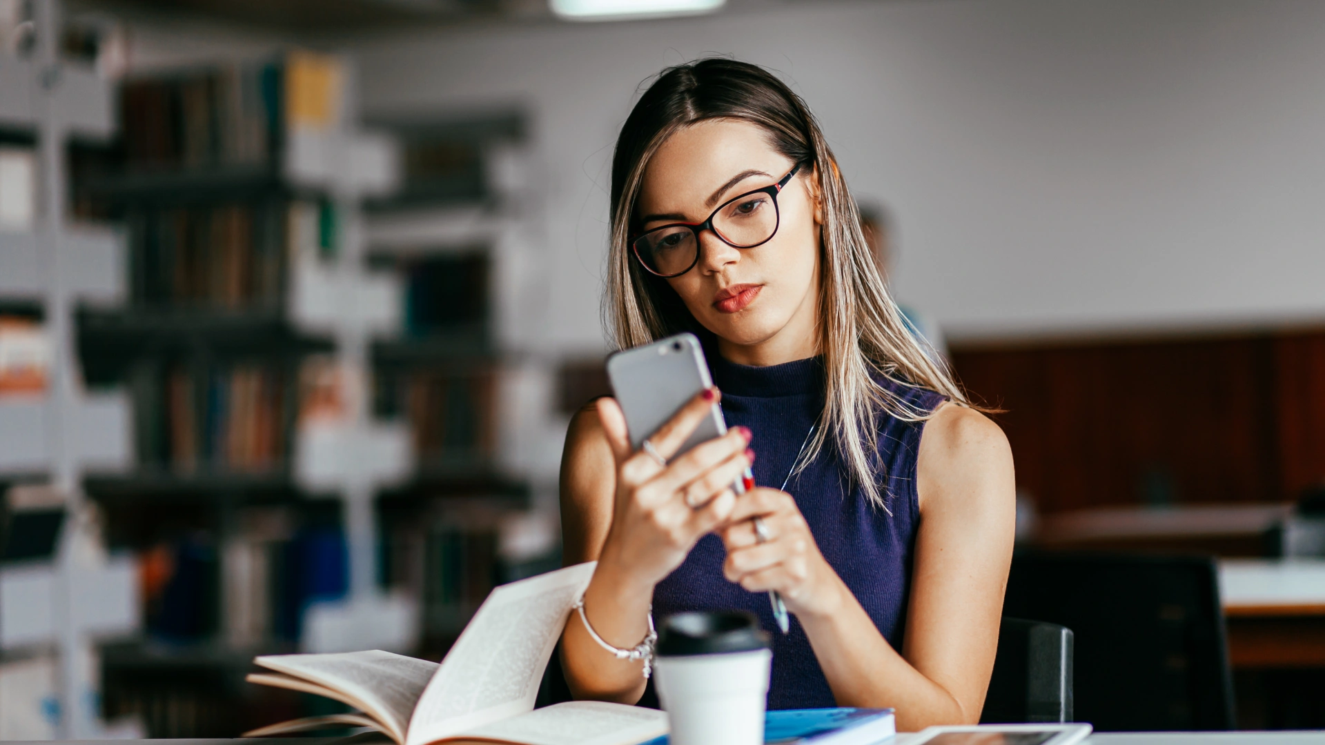 Estudante focada usando smartphone em uma biblioteca com livros e café na mesa