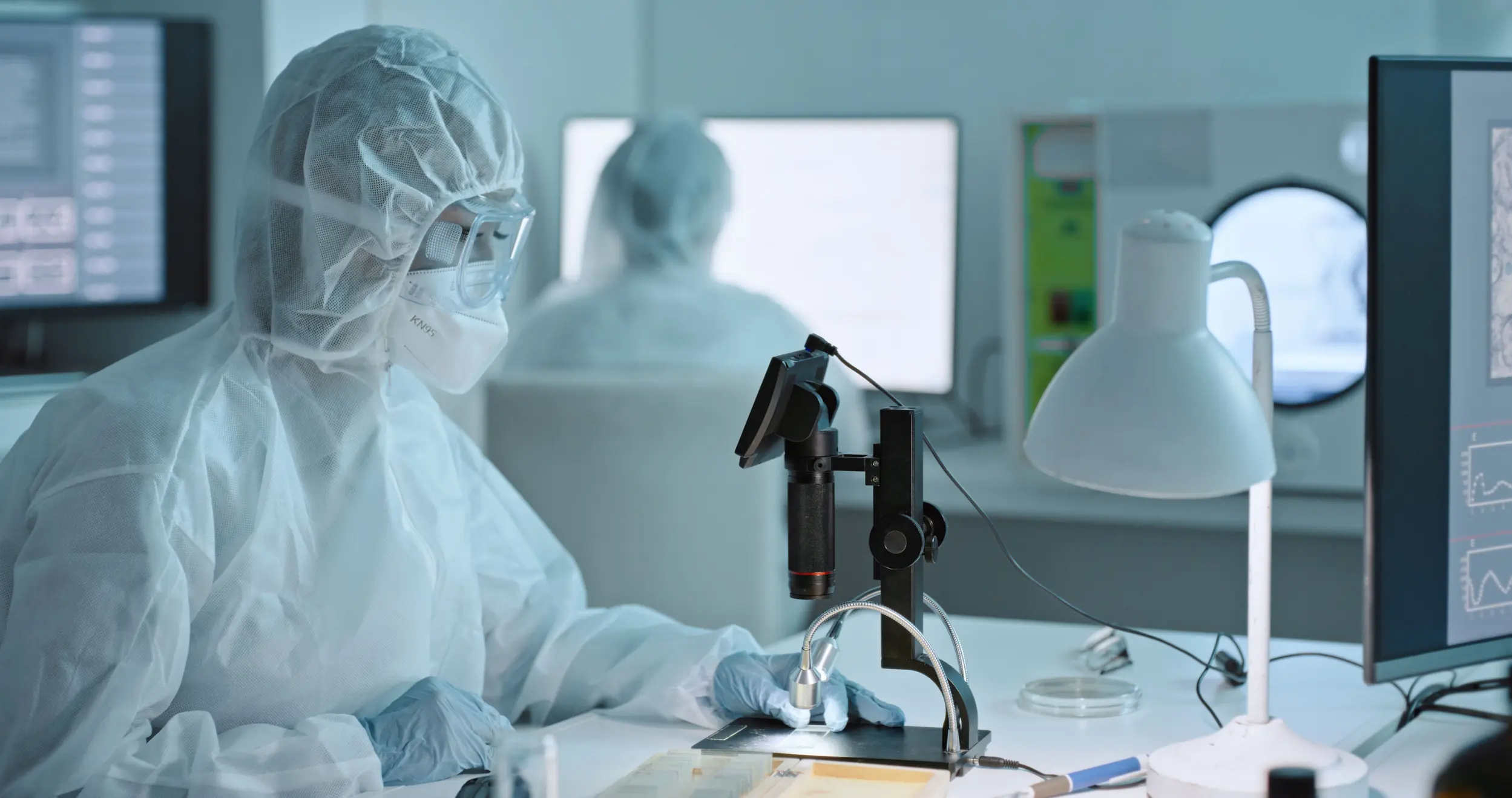 Pesquisador em laboratório vestindo traje de proteção e máscara analisando amostras ao microscópio digital.