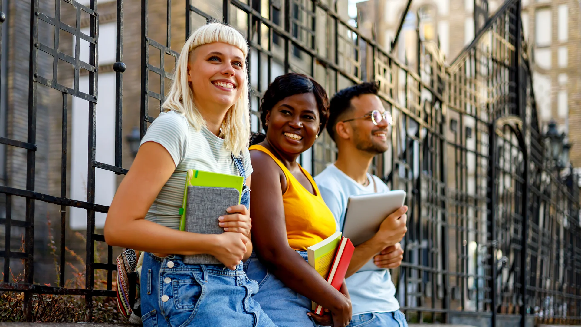 Grupo de estudantes universitários diversos sorrindo e segurando livros em frente a um portão de metal