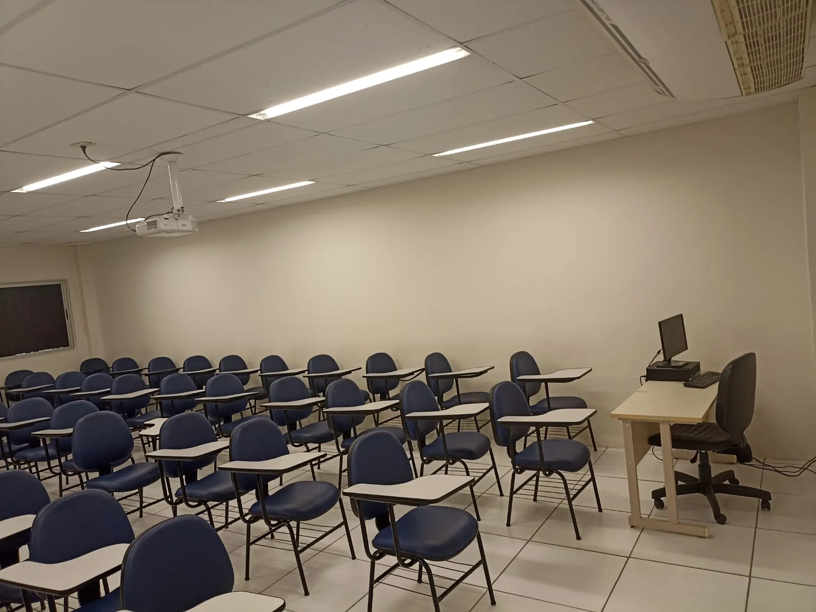Tour virtual Boa Vista UniFG-PE: sala de aula com cadeiras universitárias azuis em fileiras, projetor no teto, quadro-negro e mesa do professor com computador