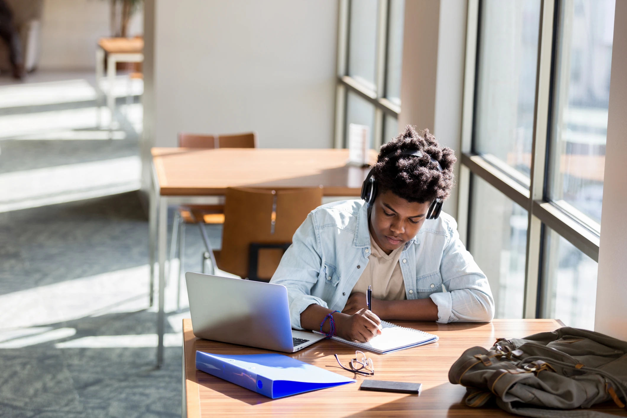 Estudante jovem negro com fones de ouvido escrevendo em um caderno em uma mesa com laptop