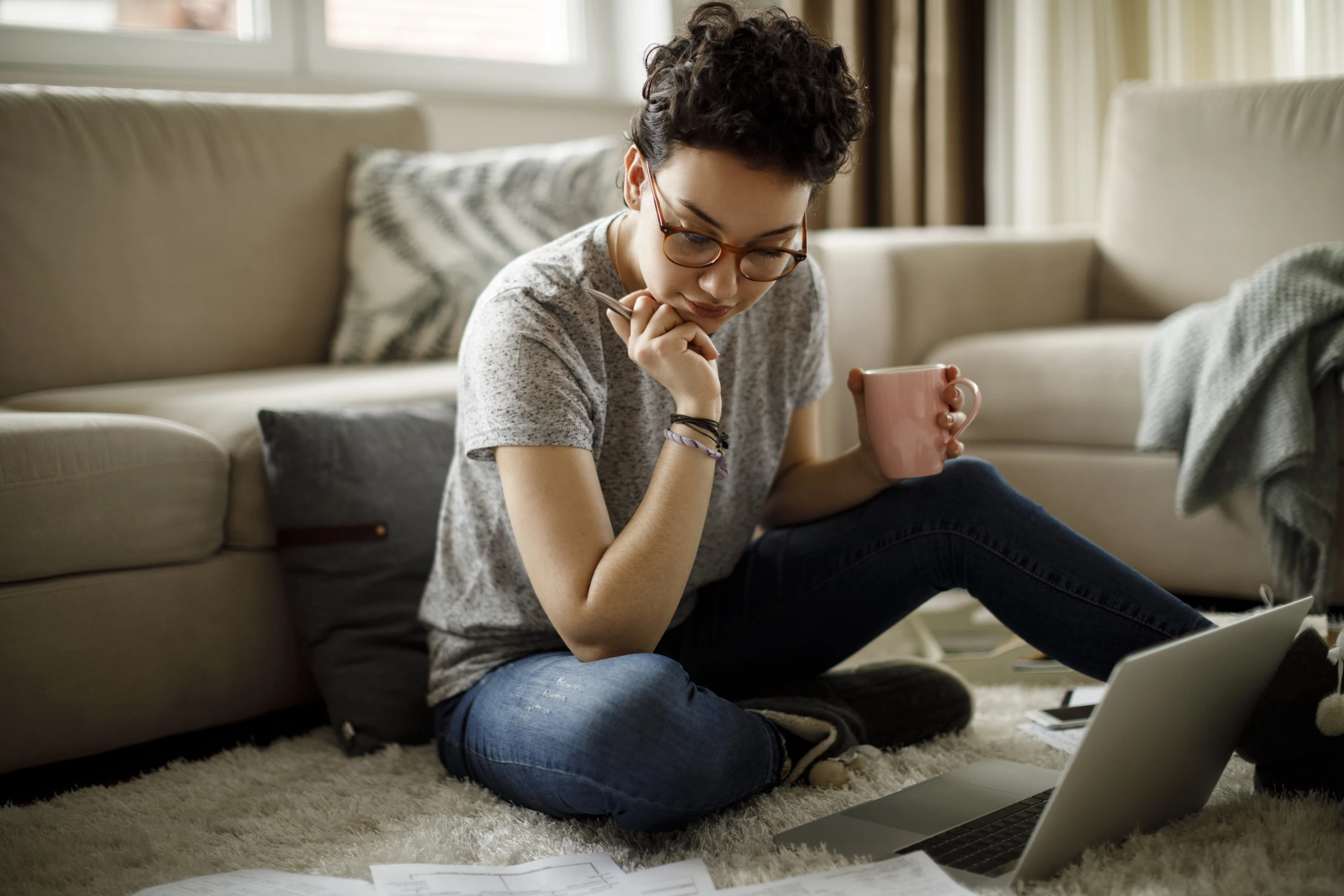 Estudante focada usando notebook e segurando uma caneca sentada no tapete da sala