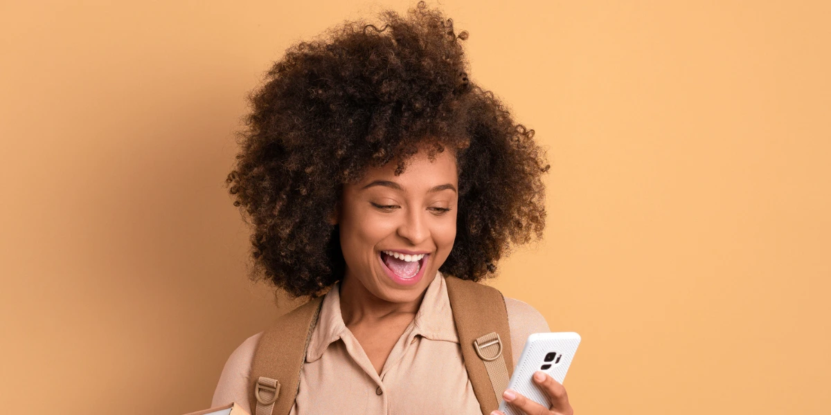 Estudante negra sorrindo ao olhar para o celular e segurando mochila